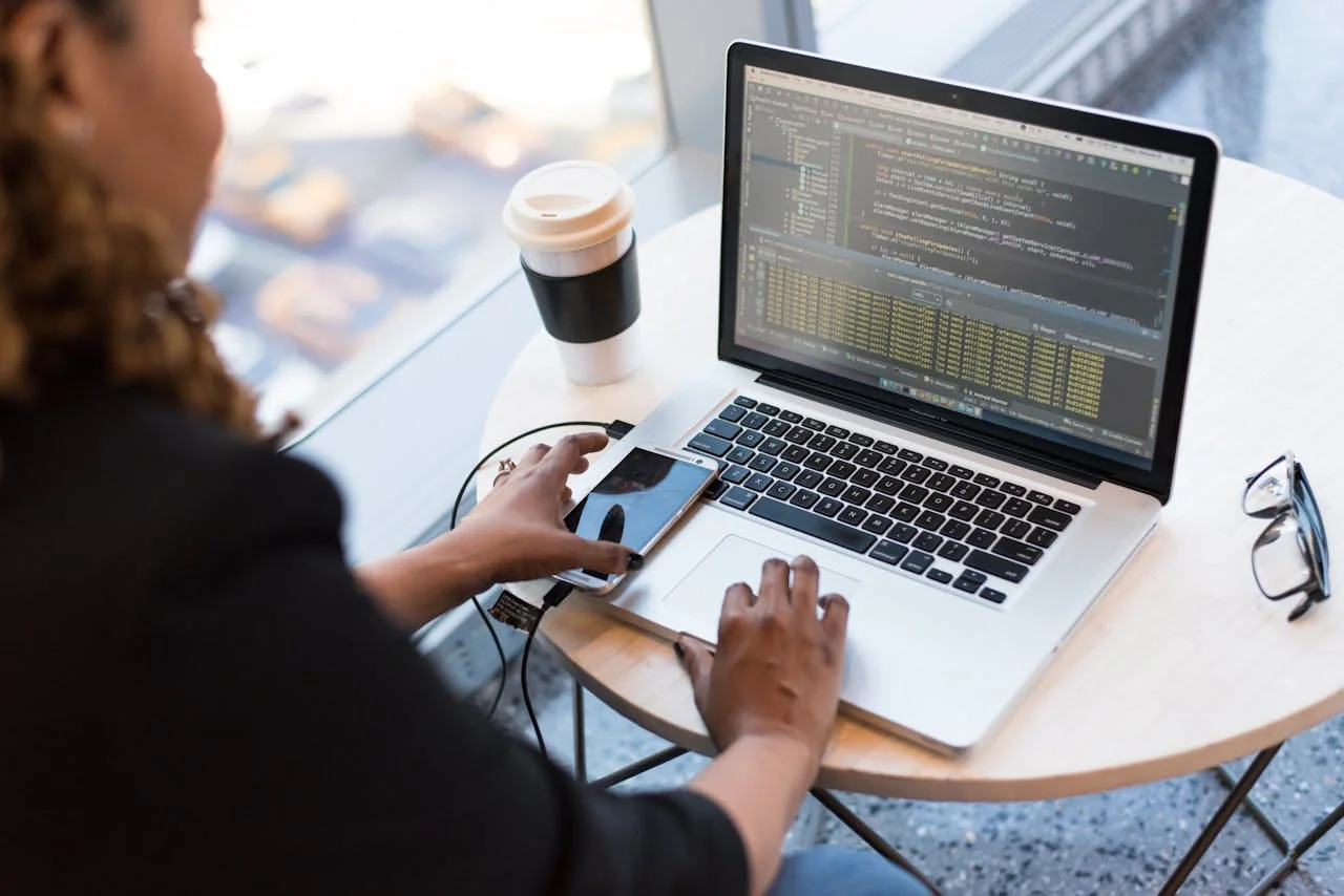 Home Black woman programming on a laptop with coffee, smartphone, and glasses on a desk in an office.